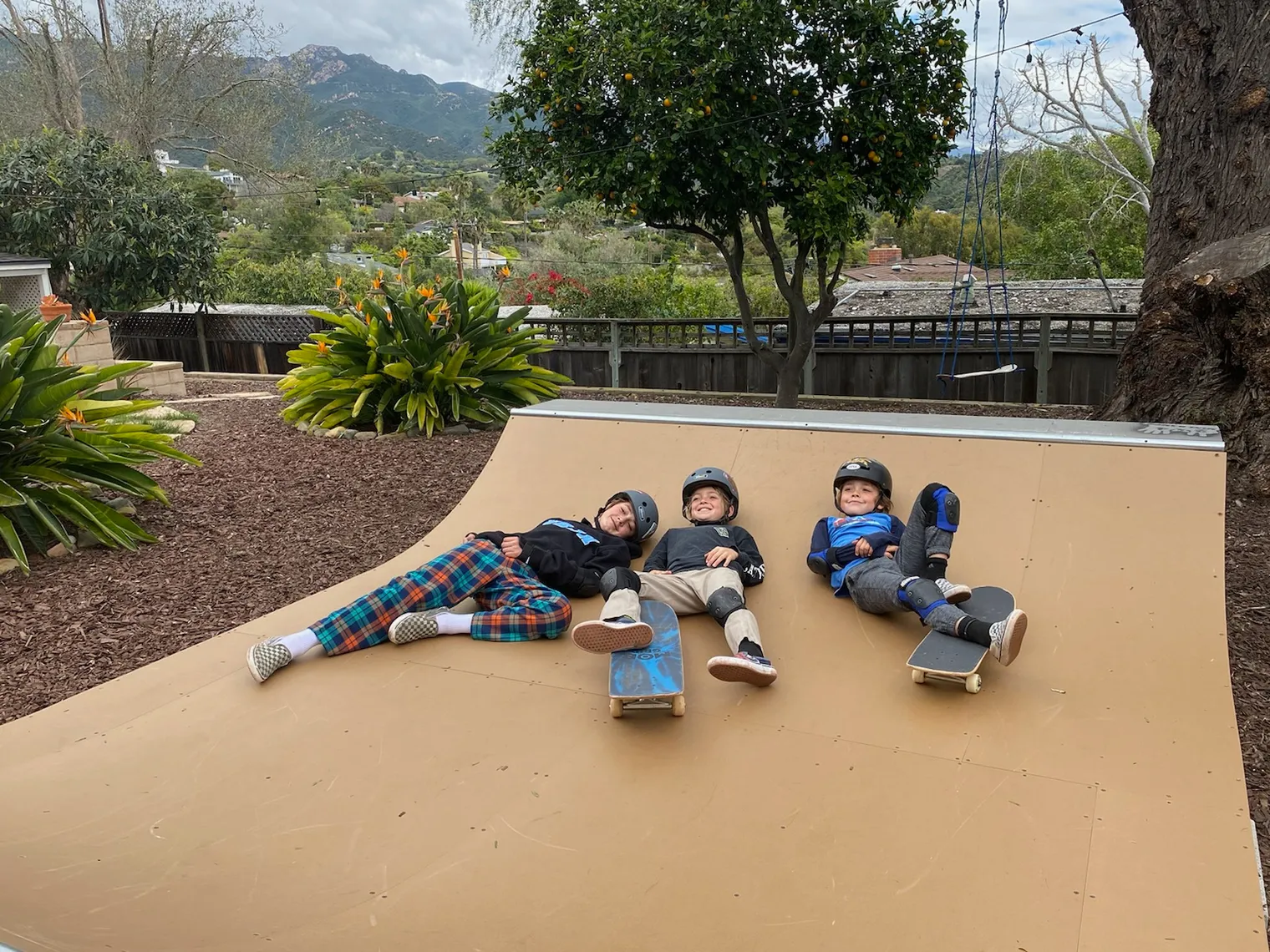 3 smiling Children in pads and helmets with skateboards laying on flat bottom of 2x4 built miniramp