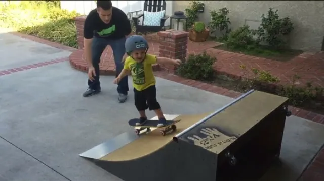 Young child in helmet riding up two foot high by four foot wide quarter pipe while dad supervises in the background
